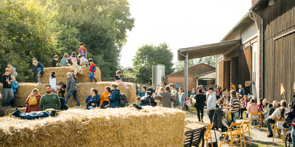 Viele Menschen, darunter Kinder, bewegen sich zwischen großen Heuballen auf einem Hofgelände vor Scheunengebäuden.